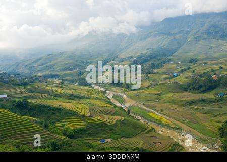Aerial view of layered rice terraces and a winding river stretching through a misty green valley near Sapa, Vietnam. Low clouds drift above the rolling hills, creating a serene and atmospheric rural landscape. Stock Photo
