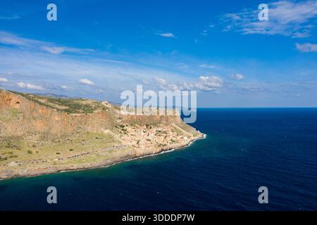 Sweeping aerial image of Monemvasia's dramatic cliffs meeting the deep blue Mediterranean Sea, with a historic village nestled at the rocky shoreline under a vibrant sky. The scene features textured stone, lush greenery, and expansive water. Stock Photo