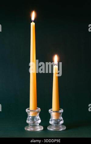 Two yellow beeswax taper candles burning in glass candle holders on dark green background Stock Photo