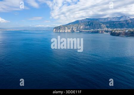 Sheer limestone cliffs rise above deep blue waters on the Bay of Naples, with distant mountains and scattered white clouds creating a striking Mediterranean panorama. The scene is defined by crisp daylight, tranquil reflections, and the natural textures of the Italian coastline. Stock Photo