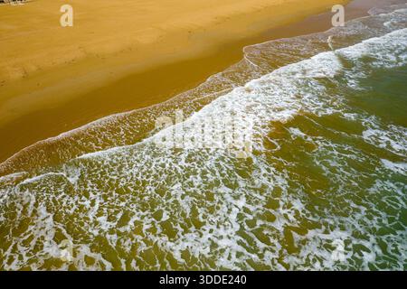 Soft waves create foamy patterns as they wash onto the expansive golden sand beach of Vieste, Italy. The aerial view highlights the natural textures and subtle color gradients of the Adriatic coast under diffused daylight. Stock Photo