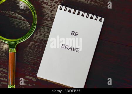 A notepad stating BE BRAVE sits beside a gold magnifying glass on a dark rustic surface. The scene inspires confidence and encourages a deeper look at Stock Photo