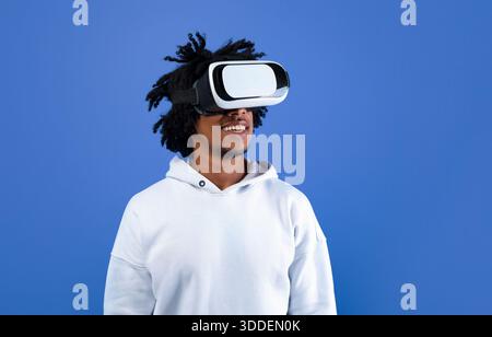 Cool African American guy wearing VR headset, using virtual reality for entertainment on blue studio background Stock Photo