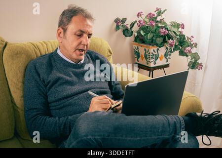 Senior man working from home, taking notes while using a laptop on sofa. Concept of remote work, concentration, mental focus, reflective thinking and Stock Photo