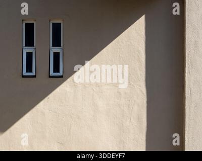 Two aluminium rectangular glass window frame on a beige stone cement wall with diagonal shadow. Stock Photo