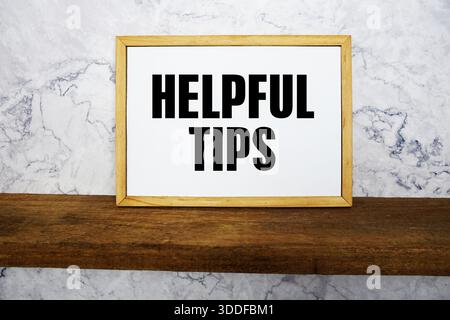 Blackboard with text "Helpful Tips" on a wooden shelf, set against a marble wall. Stock Photo