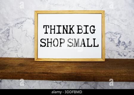 Think Big Shop Small text written on whiteboard with wooden shelf and marble background Stock Photo