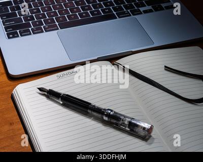An open notebook shows Sunday handwritten on the page next to a clear fountain pen and laptop on a wooden desk. Stock Photo