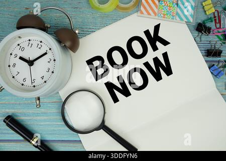 Book Now text written on notepad with alarm clock and magnifying glass flat lay on wooden background Stock Photo