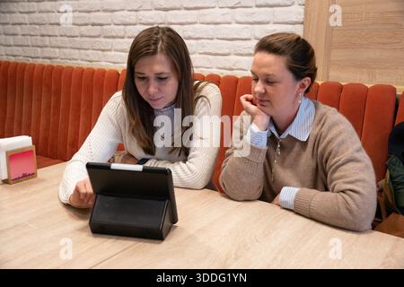 Two women sit in modern cafe using a tablet with electronic menu, focused and relaxed, with warm interior tones and a cozy atmosphere Stock Photo