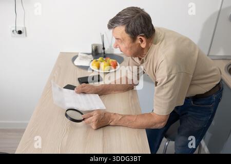 Senior male examining paperwork closely at home workspace with magnifier focused posture and everyday household setting. Stock Photo