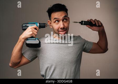 Portrait of fooling young man playing funny joke, using large cordless drill as earphone and small electric screwdriver as portable music player Stock Photo