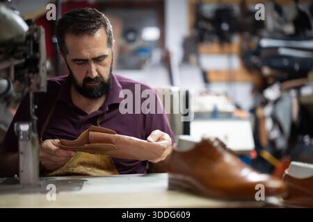 Cobbler craftsman works on leather shoes at a busy workshop, focused on stitching and finishing Stock Photo