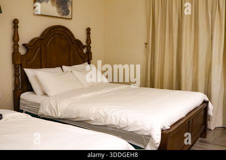 Neatly arranged bedroom with two beds, white linens, wooden headboard, beige curtains, and abstract wall art in a warm, inviting ambiance. Stock Photo
