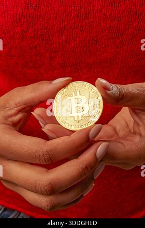 A person is holding a round coin with the Bitcoin logo in their hands. The setting is casual, and the person appears focused on the coin. Stock Photo