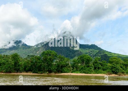White clouds float across the peaks of a lush green mountain above the Nam Ou River in northern Laos. Dense tropical forest lines the riverbank, creating a vibrant landscape beneath a bright sky. Stock Photo