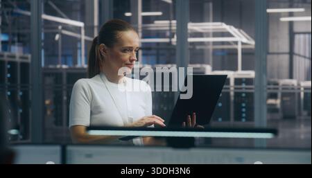 Mobile IT Management, Focused Female IT Professional Standing and Actively Working on Thin Laptop. Background Modern Server Hall. Emphasizes Mobile Enterprise Management and High Level IT Operations. Stock Photo