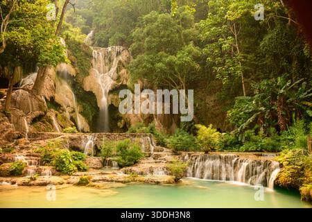 Warm sunlight bathes the layered limestone cascades and emerald pools of Kuang Si Waterfalls in northern Laos. Dense tropical forest surrounds the flowing water, highlighting lush greenery and natural textures. Stock Photo