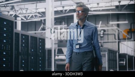 IT Executive Inspection, Senior Manager Walking in Mission Critical Server Room. Grey Haired Man in Blue Shirt and Glasses Walks While Holding Laptop Through Vast Server Room. Infrastructure Control. Stock Photo