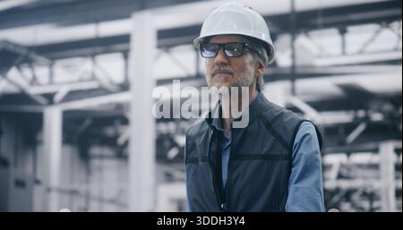 Experienced Male Executive Walking Through Modern Factory. Mature Man in Hard Hat and Vest Walking in Large, Modern Factory Setting. Concept of Industrial Leadership and on Site Decision Making. Stock Photo