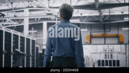 Executive Inspecting Data Center Flow. Grey Haired Man Walking and Carrying Laptop Through Enormous Scale of Data Center. Strategic Oversight and Technological Infrastructure Management. Back View. Stock Photo