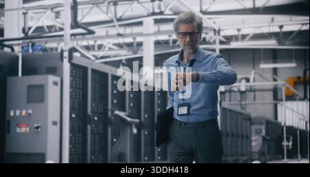 IT Executive Inspection, Senior Manager Walking in Mission Critical Server Room. Grey Haired Man in Blue Shirt and Glasses Walks While Holding Laptop Through Vast Server Room. Infrastructure Control. Stock Photo