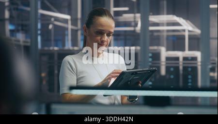 Woman, IT Specialist Stands and Uses Portable Tablet Device to Check Real Time Performance Metrics in High Security Server Hall. Concept of System Monitoring, on Site Expertise, and Data Analysis. Stock Photo