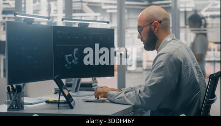 Bearded Male Engineer Programming or Developing Digital Twin, Deeply Focused on Dual Monitors Displaying Robot 3D Model on Background Modern Factory Floor. Concept of Design and Industry 4.0 Software. Stock Photo