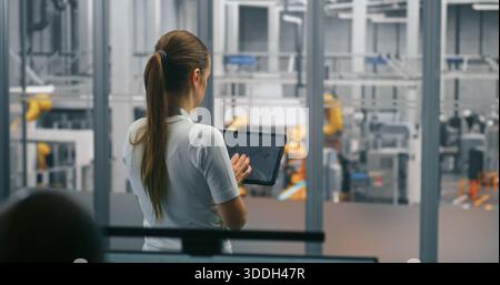 Female Engineer Managing Production Via Tablet. Woman Standing and Operating Digital Device. Clear Background Shows Automated Machinery and Robotics of Smart Factory. Mobile Control, Industry 4.0. Stock Photo