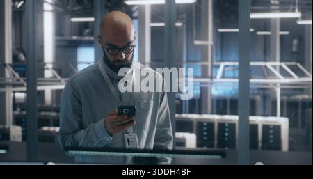 Bearded Male IT Engineer in Glasses Stands and Uses Smartphone to Check Critical Notifications or System Status Large Scale of Data Center. Emphasizes Mobile Management and Operational Urgency. Stock Photo