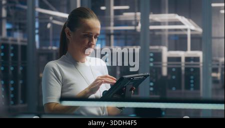 Woman, IT Specialist Stands and Uses Portable Tablet Device to Check Real Time Performance Metrics in High Security Server Hall. Concept of System Monitoring, on Site Expertise, and Data Analysis. Stock Photo