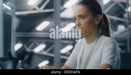 Focused Female IT Engineer Concentrating on System Monitoring Workstation. Background Blue Lit, High Tech Environment of Data Center Control Room. Critical Analysis and Professional Dedication. Stock Photo