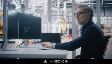 Focused Male Professional Programs Robotic Equipment While Tracking Operational Metrics on Dual Monitor Desk in Modern Factory Setting. Concept of Industrial Automation and Production Strategy. Stock Photo