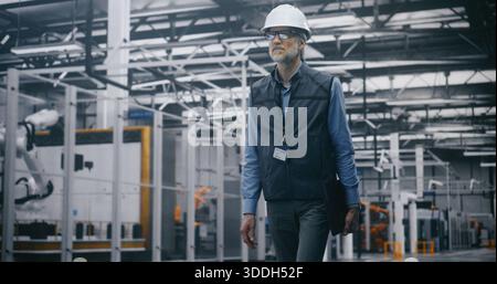 Grey Haired Male Senior Engineer in Safety Vest and Helmet Walks and Holds Laptop, Conducting Safety Audit in Modern Robotics Factory. Concept of Industrial Safety, Audit and Expert Supervision. Stock Photo