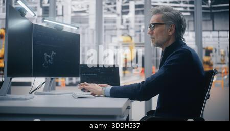 Focused Male Professional Programs Robotic Equipment While Tracking Operational Metrics on Dual Monitor Desk in Modern Factory Setting. Concept of Industrial Automation and Production Strategy. Stock Photo