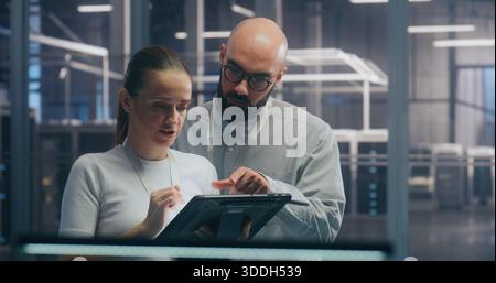 Male and Female IT Engineer Team Collaborating and Discussing Data Displayed on Digital Tablet, Standing in Secure, High Tech Environment of Data Center. Concept Teamwork and Mobile System Diagnostic. Stock Photo