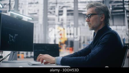 Senior Engineer Typing Dual Screen Workstation, Monitoring 3D Robotic Model on Screen and System Performance With Robotic Arms in Background. Concept of Digital Twin Technology and Real Time Control. Stock Photo