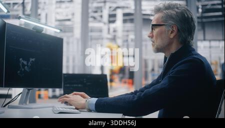 Senior Engineer Typing Dual Screen Workstation, Monitoring 3D Robotic Model on Screen and System Performance With Robotic Arms in Background. Concept of Digital Twin Technology and Real Time Control. Stock Photo