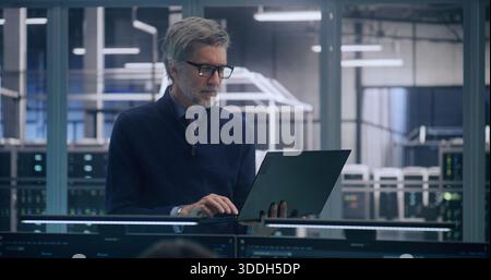Focused Senior Manager in Formal Business Casual Wear Stands And Works, Uses Laptop to Analyze Data in Large, Mission Critical Server Room. Concept of Strategic Analysis and High Level IT Management. Stock Photo