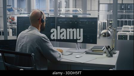 Cloud System Monitoring, Engineer on Headset Analyzing Performance Metrics, Working Two Monitors and Tablet. Screens Display Complex Cloud System Metrics and Graphs on Backdrop of Large Server Hall. Stock Photo