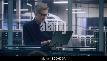 Focused Senior Manager in Formal Business Casual Wear Stands And Works, Uses Laptop to Analyze Data in Large, Mission Critical Server Room. Concept of Strategic Analysis and High Level IT Management. Stock Photo