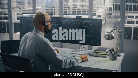 Specialist Coordinating Data Center Operations Remotely. IT Specialist Speaking on Headset, Actively Typing at Desk With Dual Monitors in High Security Data Center. Concept of Focused Data Analysis. Stock Photo
