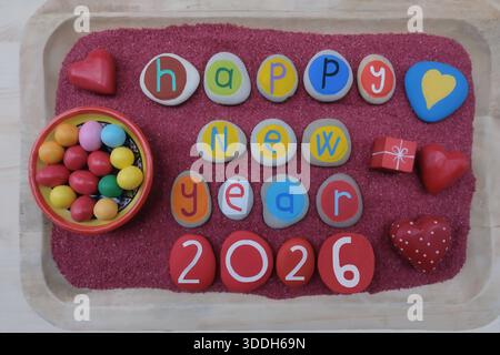 Happy New Year 2026 celebrated with hand painted stones for a healthy future full of love and sweets over red sand and a wooden board Stock Photo