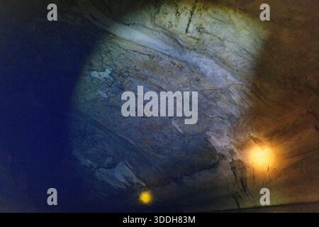 Bands of blue and amber artificial light illuminate the textured limestone ceiling inside Konglor Cave in central Laos. The interplay of colors highlights mineral patterns and the rugged surface of this vast subterranean cavern. Stock Photo