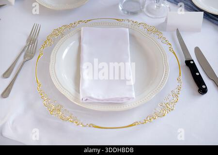 Elegant formal table setting featuring a glass plate with ornate gold details placed on a crisp white tablecloth with a neatly folded napkin on top Stock Photo