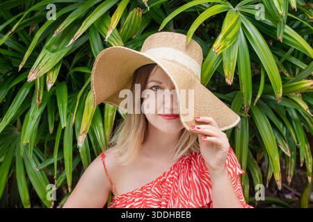 Young woman with soft blonde hair posing in front of dense tropical greenery, wearing a large straw hat and a red patterned summer dress Stock Photo