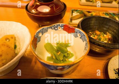 Lightly seasoned green vegetables are served in a colorful porcelain bowl, surrounded by assorted Japanese breakfast dishes on a wooden table. Warm lighting enhances the inviting textures and vibrant presentation. Stock Photo