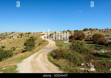 Curving dirt road cuts through a sunlit plateau with scattered bushes, rocky ground, and vibrant blue sky, evoking a peaceful countryside atmosphere near Matera, Italy. Stock Photo