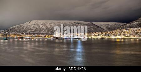Winter night panorama of Tromso, Norway with glowing city lights, bridge and Arctic Cathedral across a fjord beneath snow-covered mountains. Stock Photo