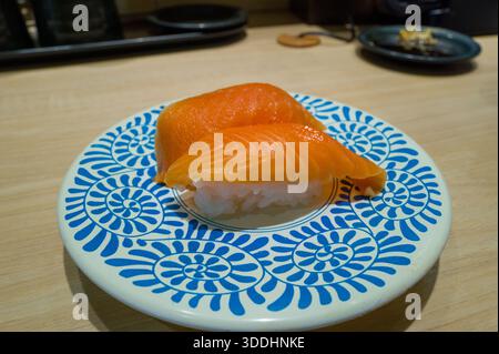 A pair of salmon nigiri sushi is presented on a white plate with bold blue geometric patterns, set on a light wooden counter in a contemporary Osaka sushi restaurant. Stock Photo